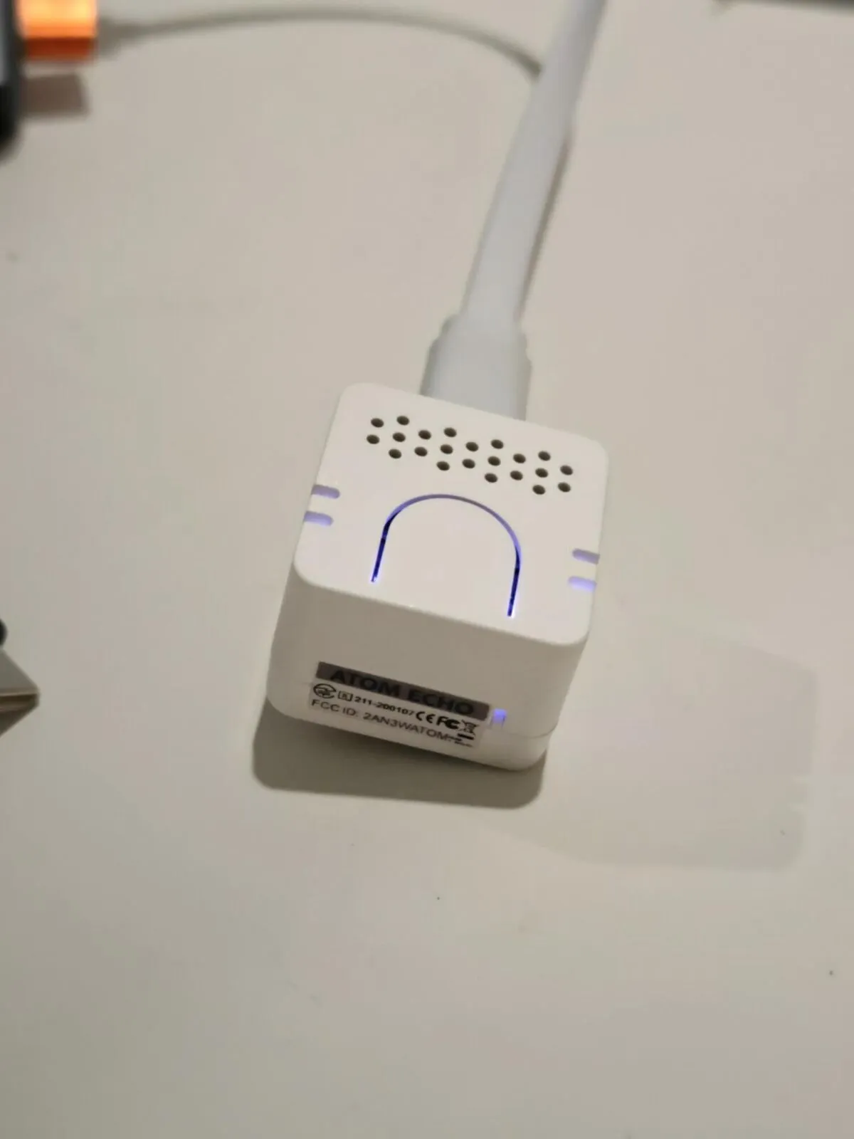 The Atom Echo from above: a white cube with a grille of small holes, a U-shaped slot lit blue, and a label on its side reading ATOM ECHO